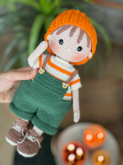 A handmade amigurumi crochet Boy With Pumpkin Hat, green overalls, and striped shirt. This customizable doll comes with a name label, perfect for fall or as an autumn gift.