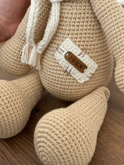 A handmade beige teddy bear toy crafted with the amigurumi technique, featuring a soft texture, a personalized leather name patch, and a cute, cuddly design.