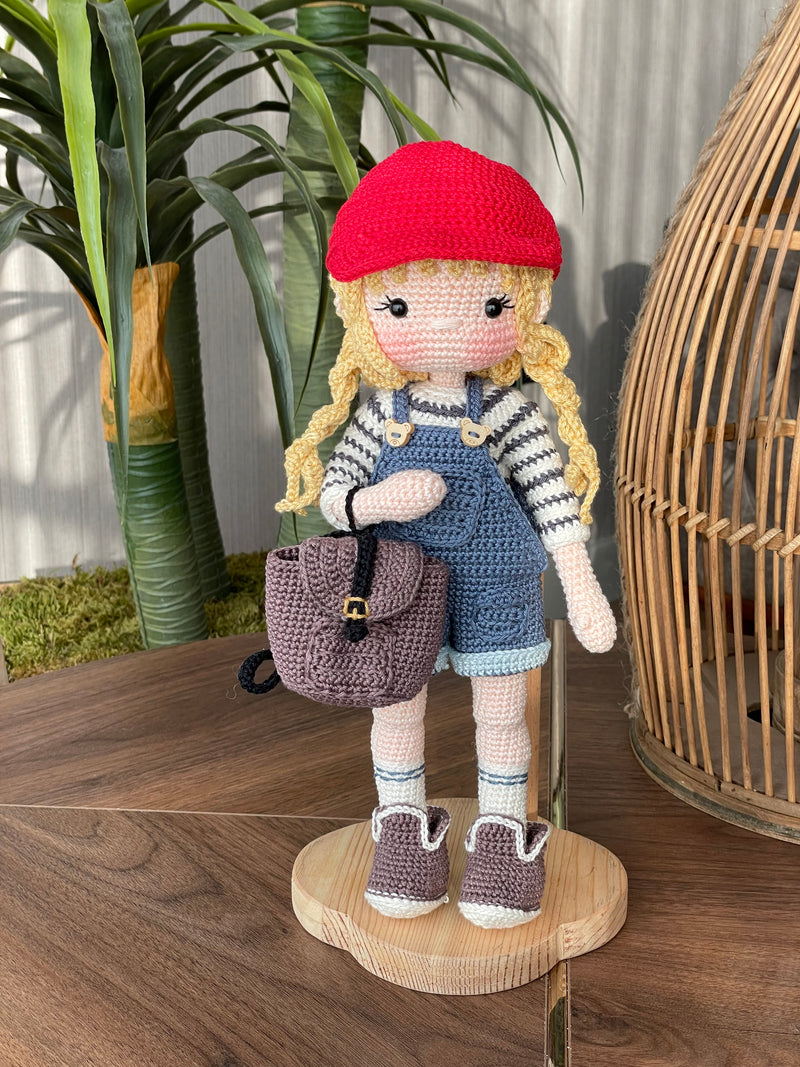 Handmade crocheted Mila Doll with a red cap, blue overalls, and a striped shirt, holding a small backpack, made with 100% cotton yarn and customizable with a name on a leather label.