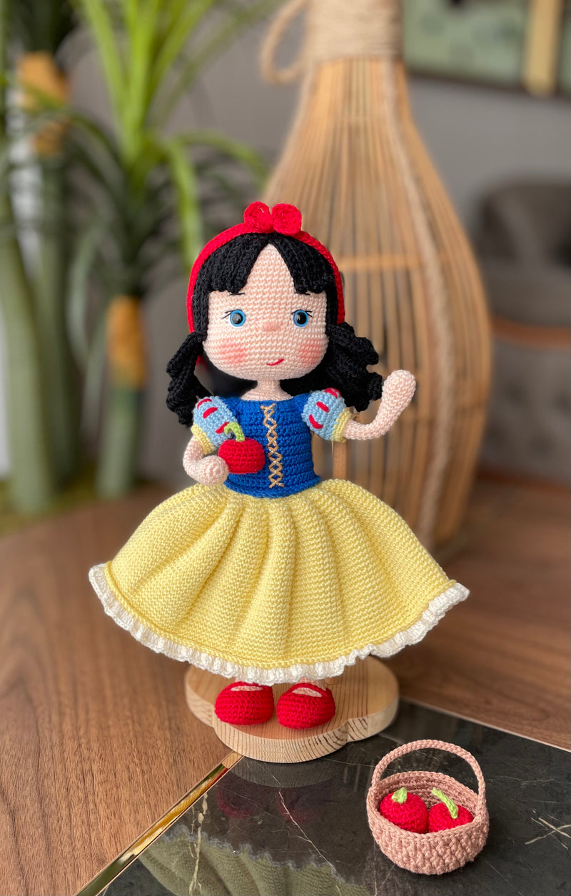 Handmade Snow White Doll wearing a classic yellow dress with a blue bodice, holding a basket of apples, made with 100% cotton yarn, customizable with a name on a leather label.