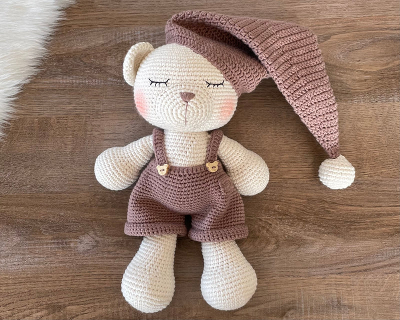 Handmade crocheted Sleeping Teddy Bear Toy in soft, neutral tones with a cozy hat and overalls, customizable with a name on a leather label. Perfect for bedtime cuddles and nursery decor.