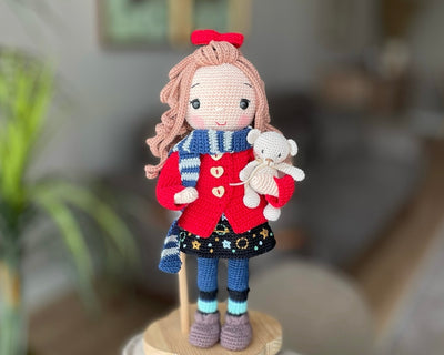 Handmade crochet Idealistic Girl Doll with a red coat, blue scarf, and holding a small teddy bear. This amigurumi doll is customizable with a name on a leather label, perfect for personalized gifts.