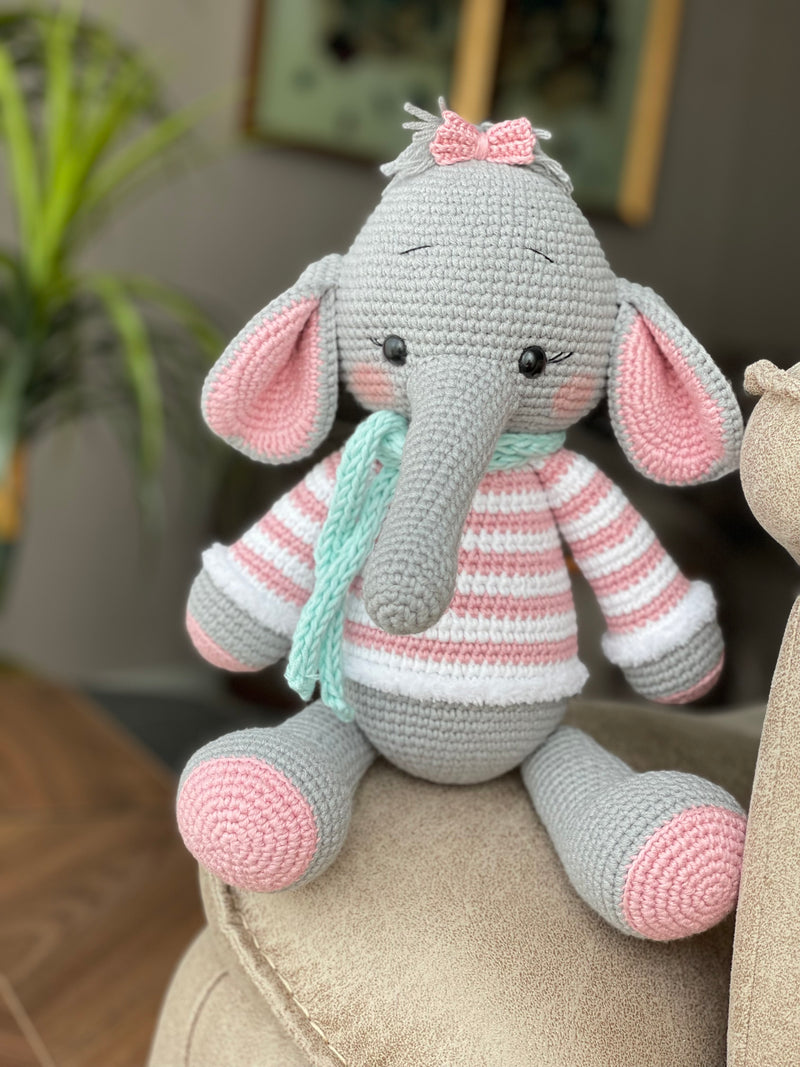 A handmade crochet elephant toy featuring a soft, cuddly design with large pink ears, pink striped sweater, and a mint green scarf, made using the amigurumi technique and personalized with a custom name on a leather label.