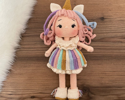 A handmade crocheted Unicorn Girl Doll with pastel rainbow dress, pink curly hair, and a golden horn. This amigurumi toy is customizable with a name on a leather label, perfect for nursery decor and personalized gifts.