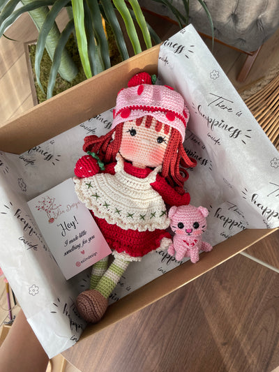 Handmade strawberry shortcake crochet doll with red hair, wearing a strawberry-themed outfit. Features a pink hat with red polka dots, red dress with white apron, and striped stockings. Perfect for children and collectors. Inside the gift box