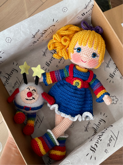 A handmade Rainbow Brite Girl Doll with vibrant yellow hair, holding Twink, a colorful star plush toy. The doll is crocheted using the amigurumi technique and personalized with a custom name on a leather label. Lying inside a gift box.