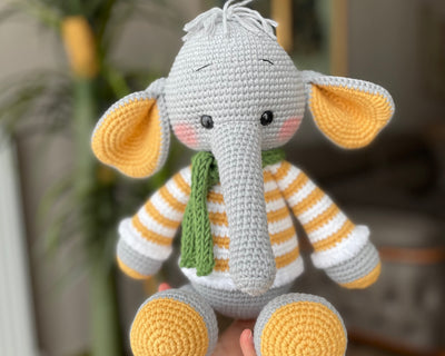 A handmade crochet elephant toy featuring a soft, cuddly design with large yellow ears, yellow striped sweater, and a green scarf, made using the amigurumi technique and personalized with a custom name on a leather label.