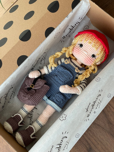 Handmade crocheted Mila Doll with a red cap, blue overalls, and a striped shirt, holding a small backpack, made with 100% cotton yarn and customizable with a name on a leather label. Lying inside a gift box.