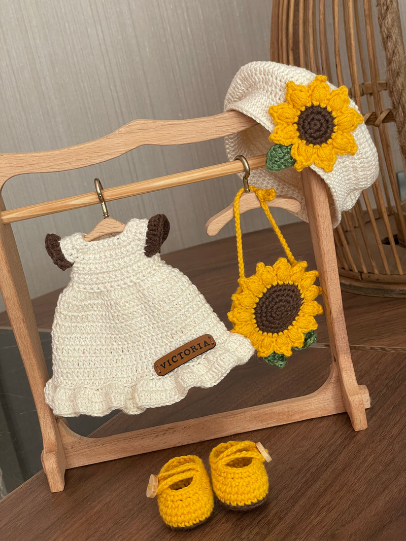 A handmade Sunflower Girl Doll with dark brown hair, wearing a white dress adorned with sunflowers, a matching sunflower beret, and yellow shoes, crocheted using the amigurumi technique and personalized with a custom name on a leather label.