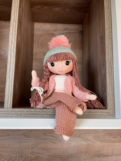 A handcrafted Pink Cardigan Cool Girl Doll with long brown hair, wearing a stylish pink cardigan and beanie, crocheted using the amigurumi technique, and personalized with a custom name on a leather label.