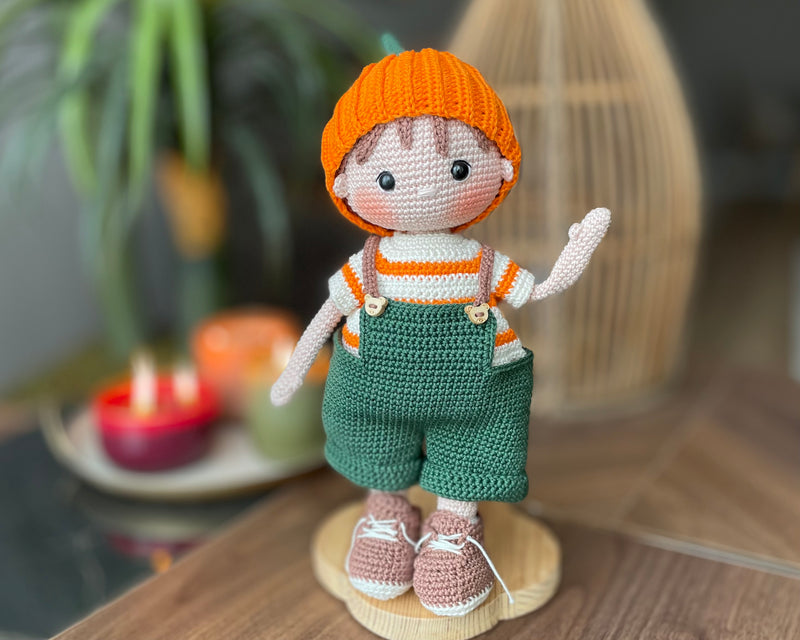 A handmade amigurumi crochet Boy With Pumpkin Hat, green overalls, and striped shirt. This customizable doll comes with a name label, perfect for fall or as an autumn gift.