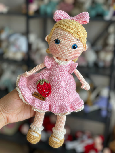 Handmade Pink Nina Doll with blonde hair, wearing a pink dress with a strawberry detail, made with 100% cotton yarn, customizable with a name on a leather label.