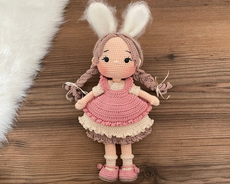 A handmade crocheted Rabbit Girl Doll with a pink dress, braided hair, and soft bunny ears. This amigurumi toy is customizable with a name on a leather label, making it a perfect choice for nursery decor and personalized gifts.