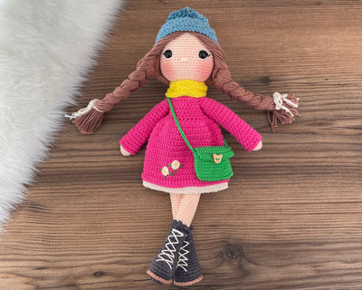 Handmade crocheted Magenta Winter Girl Doll featuring a magenta dress, flower detail, and a cute braided hairstyle. Customizable with a name on a leather label, made using amigurumi techniques. Perfect as a personalized gift or nursery decor.