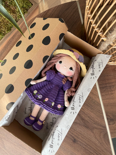 Handmade crocheted Purple Rose Girl Doll with long brown hair, wearing a purple dress with floral accents and a yellow hat. The doll is customizable with a name on a leather label, made using amigurumi techniques. Lying inside a gift box. Perfect gift for