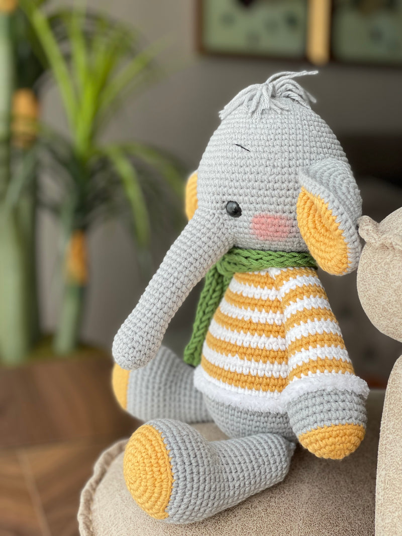 A handmade crochet elephant toy featuring a soft, cuddly design with large yellow ears, yellow striped sweater, and a green scarf, made using the amigurumi technique and personalized with a custom name on a leather label.