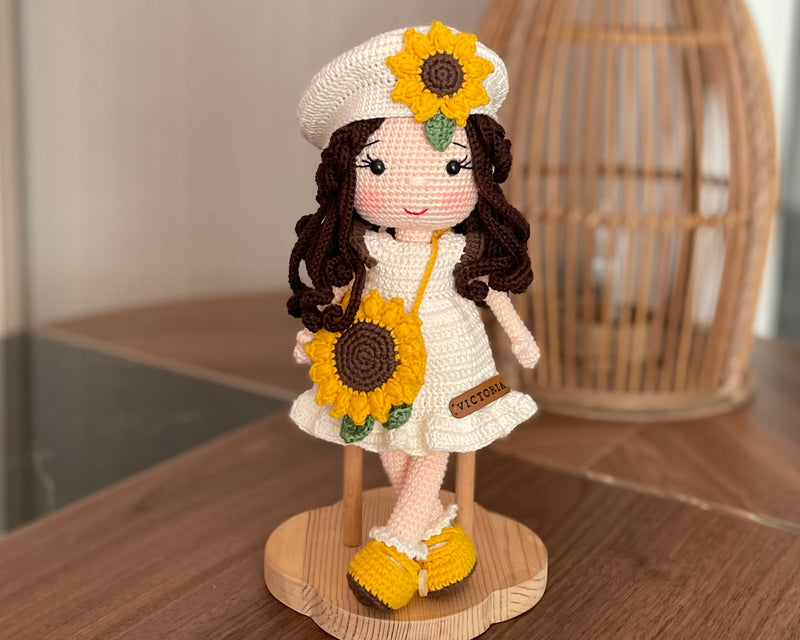 A handmade Sunflower Girl Doll with dark brown hair, wearing a white dress adorned with sunflowers, a matching sunflower beret, and yellow shoes, crocheted using the amigurumi technique and personalized with a custom name on a leather label.