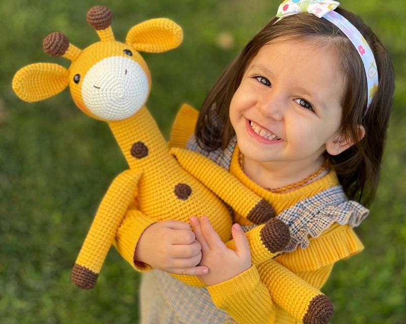 A cute handmade crocheted yellow giraffe toy held by a smiling child, made using the amigurumi technique, with customizable name on a leather label. Perfect for baby showers and personalized gifts.