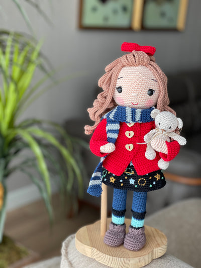 Handmade crochet Idealistic Girl Doll with a red coat, blue scarf, and holding a small teddy bear. This amigurumi doll is customizable with a name on a leather label, perfect for personalized gifts.