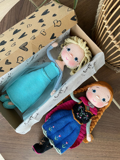 Handmade crocheted amigurumi dolls of Elsa and Anna, inspired by Frozen. Detailed with colorful outfits, perfect for custom name personalization or gifting. Lying inside a gift box.