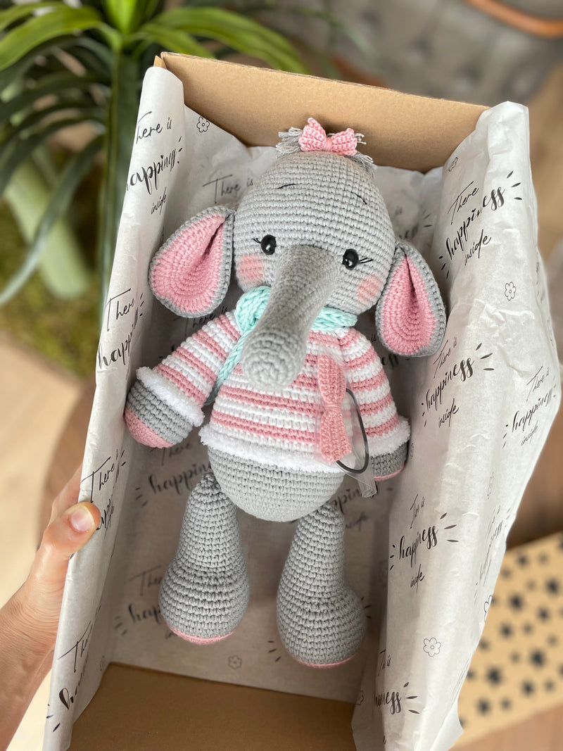 A handmade crochet elephant toy featuring a soft, cuddly design with large pink ears, pink striped sweater, and a mint green scarf, made using the amigurumi technique and personalized with a custom name on a leather label. Lying inside a gift box.