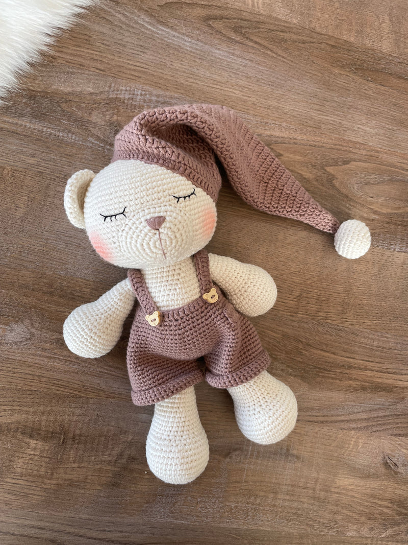 Handmade crocheted Sleeping Teddy Bear Toy in soft, neutral tones with a cozy hat and overalls, customizable with a name on a leather label. Perfect for bedtime cuddles and nursery decor.