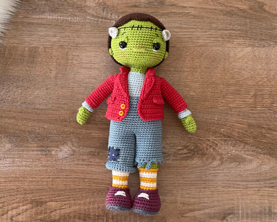 A handmade crochet Frankenstein toy featuring green skin, a red jacket, and patchwork pants. The doll is made using the amigurumi technique and is personalized with a custom name on a leather label.