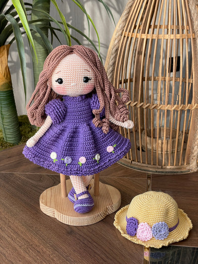 Handmade crocheted Purple Rose Girl Doll with long brown hair, wearing a purple dress with floral accents and a yellow hat. The doll is customizable with a name on a leather label, made using amigurumi techniques. Perfect gift for kids or room decor.