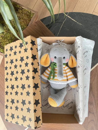 A handmade crochet elephant toy featuring a soft, cuddly design with large yellow ears, yellow striped sweater, and a green scarf, made using the amigurumi technique and personalized with a custom name on a leather label. Lying inside a gift box.