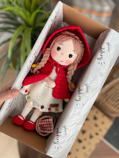 Handmade crochet Little Red Riding Hood doll, inspired by the classic fairytale, featuring a red cape, basket, and customizable name on a leather label. Lying inside a gift box. Perfect for kids and collectors.