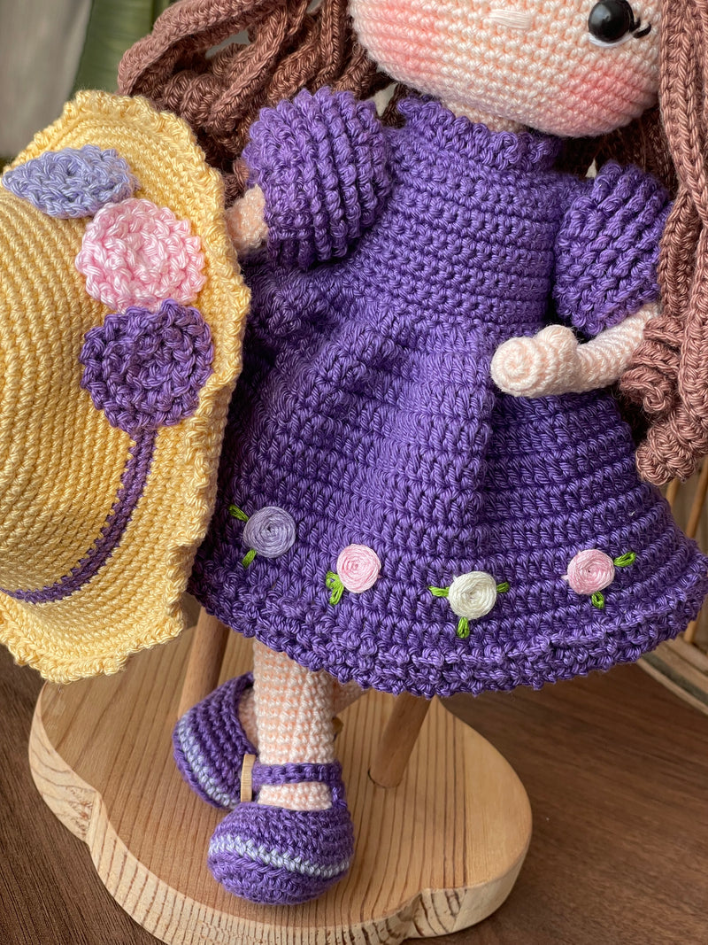 Handmade crocheted Purple Rose Girl Doll with long brown hair, wearing a purple dress with floral accents and a yellow hat. The doll is customizable with a name on a leather label, made using amigurumi techniques. Perfect gift for kids or room decor.