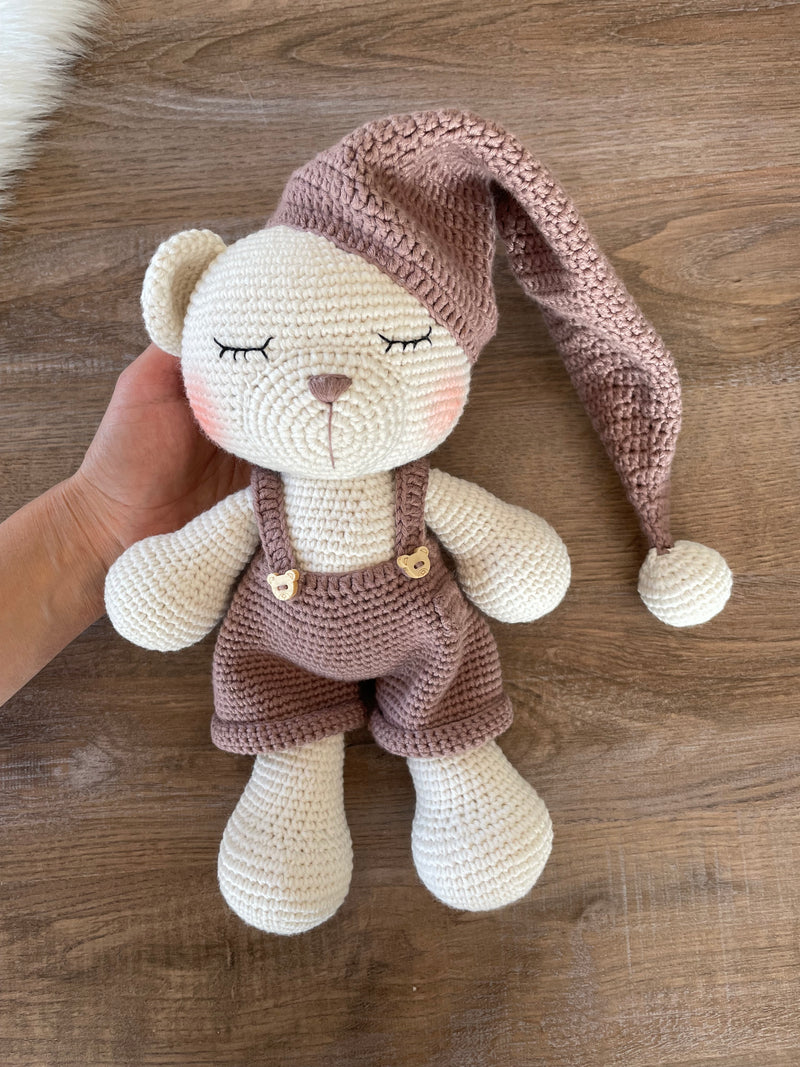 Handmade crocheted Sleeping Teddy Bear Toy in soft, neutral tones with a cozy hat and overalls, customizable with a name on a leather label. Perfect for bedtime cuddles and nursery decor.