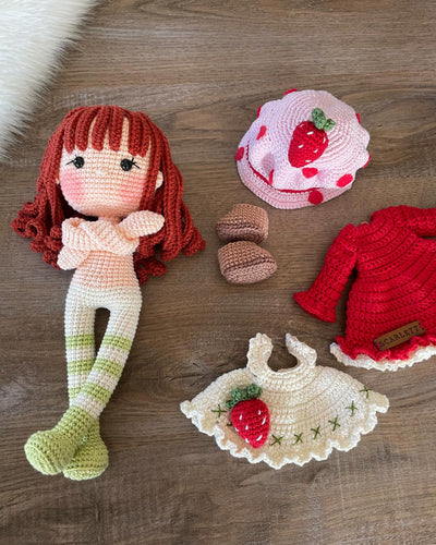 Handmade strawberry shortcake crochet doll with red hair, wearing a strawberry-themed outfit. Features a pink hat with red polka dots, red dress with white apron, and striped stockings. Perfect for children and collectors. Lying on a wooden surface.