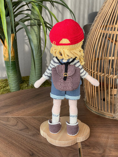 Handmade crocheted Mila Doll with a red cap, blue overalls, and a striped shirt, holding a small backpack, made with 100% cotton yarn and customizable with a name on a leather label.