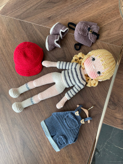 Handmade crocheted Mila Doll with a red cap, blue overalls, and a striped shirt, holding a small backpack, made with 100% cotton yarn and customizable with a name on a leather label.