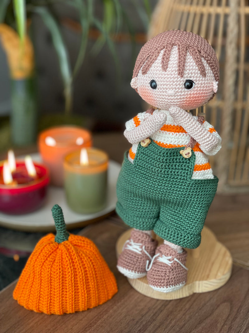 A handmade amigurumi crochet Boy With Pumpkin Hat, green overalls, and striped shirt. This customizable doll comes with a name label, perfect for fall or as an autumn gift.