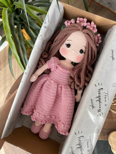 Handmade pink princess girl doll, crocheted using amigurumi technique. Features a beautiful dress and flower crown, customizable with a name on a leather label. Lying inside a gift box. Perfect for kids and collectors.