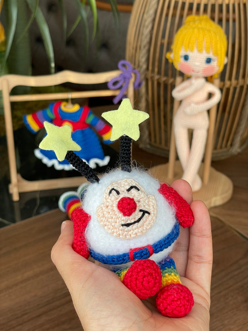 A handmade Rainbow Brite Girl Doll with vibrant yellow hair, holding Twink, a colorful star plush toy. The doll is crocheted using the amigurumi technique and personalized with a custom name on a leather label.