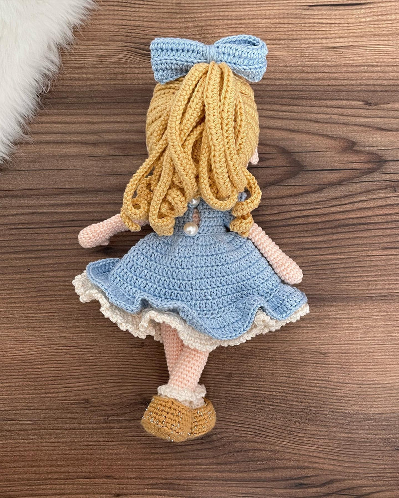 Handmade Blue Nina Doll with blonde hair, wearing a blue dress and a matching bow, made with 100% cotton yarn, customizable with a name on a leather label.