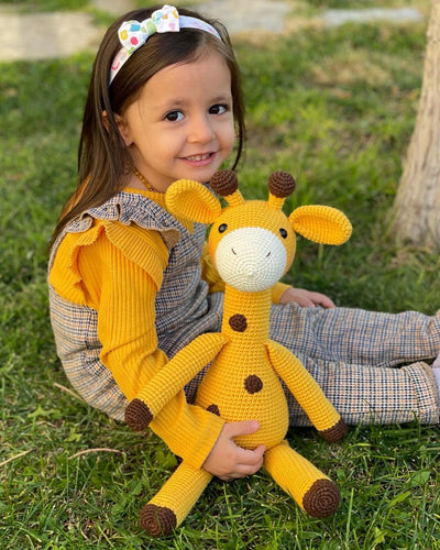 A cute handmade crocheted yellow giraffe toy held by a smiling child, made using the amigurumi technique, with customizable name on a leather label. Perfect for baby showers and personalized gifts.