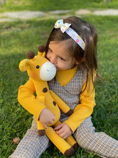 A cute handmade crocheted yellow giraffe toy held by a smiling child, made using the amigurumi technique, with customizable name on a leather label. Perfect for baby showers and personalized gifts.