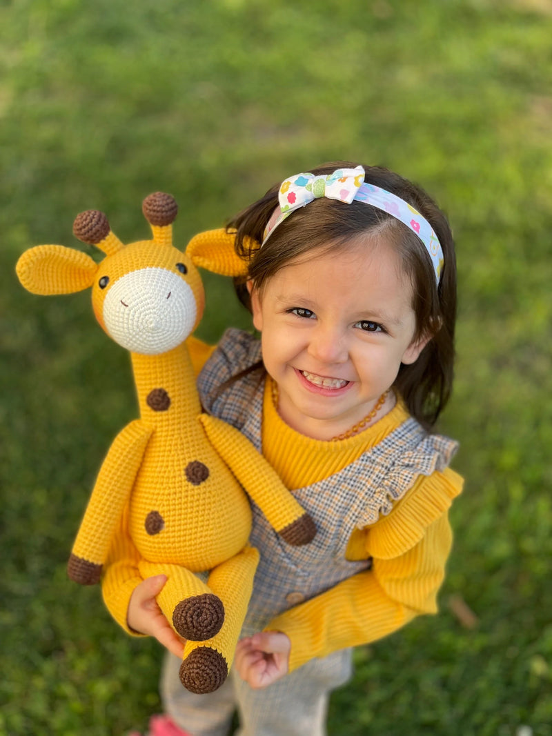 A cute handmade crocheted yellow giraffe toy held by a smiling child, made using the amigurumi technique, with customizable name on a leather label. Perfect for baby showers and personalized gifts.