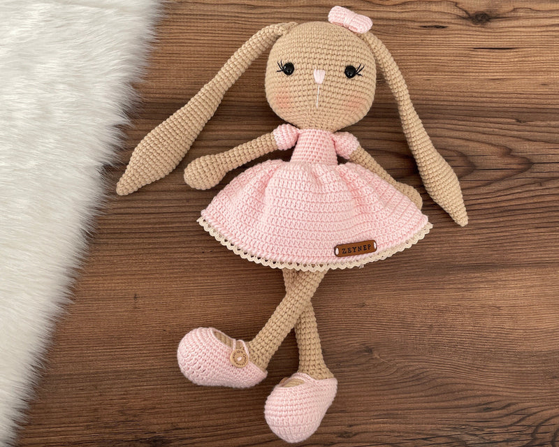 Handmade brown skin bunny rabbit toy crocheted using the amigurumi technique. Features a soft pink dress and a customizable name on a leather label. Perfect for baby gifts and nursery decor.