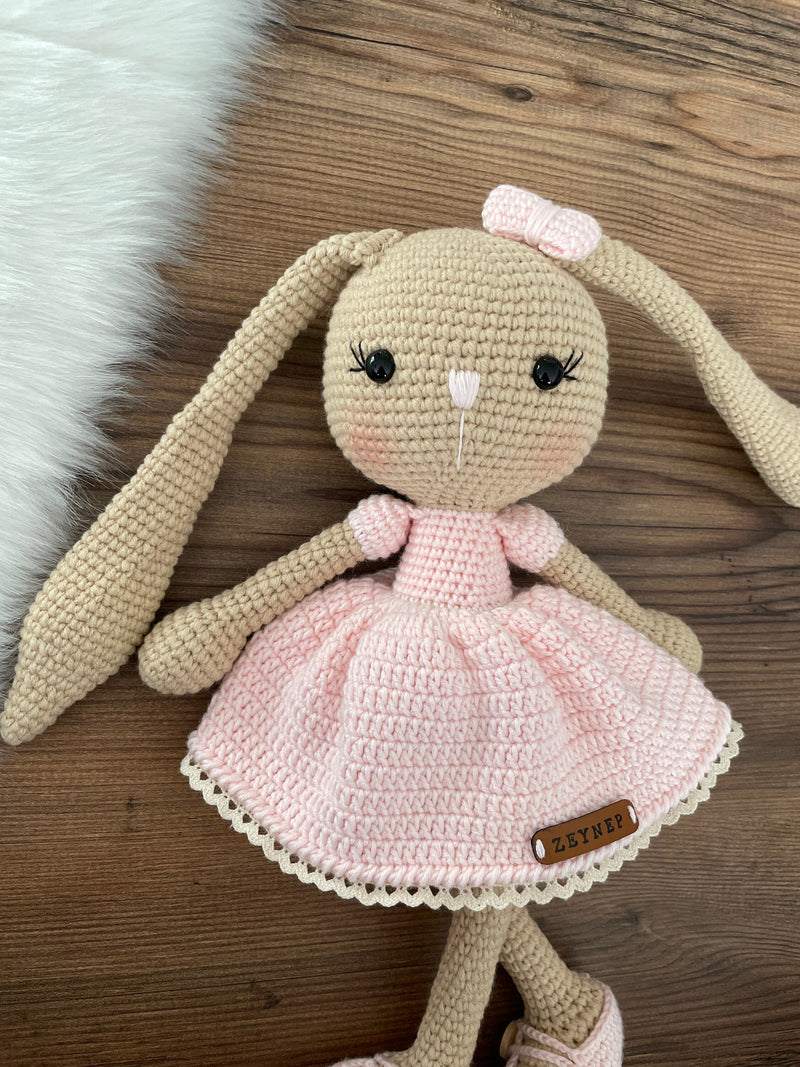 Handmade brown skin bunny rabbit toy crocheted using the amigurumi technique. Features a soft pink dress and a customizable name on a leather label. Perfect for baby gifts and nursery decor.