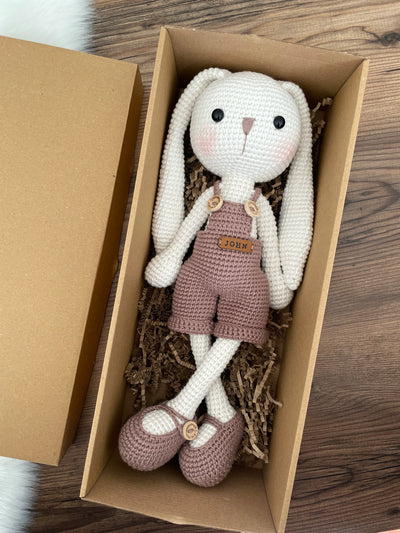 Handmade mink color overall bunny rabbit toy, crocheted using amigurumi technique with long ears and a custom name on a leather label, lying inside a gift box, perfect for gifts.