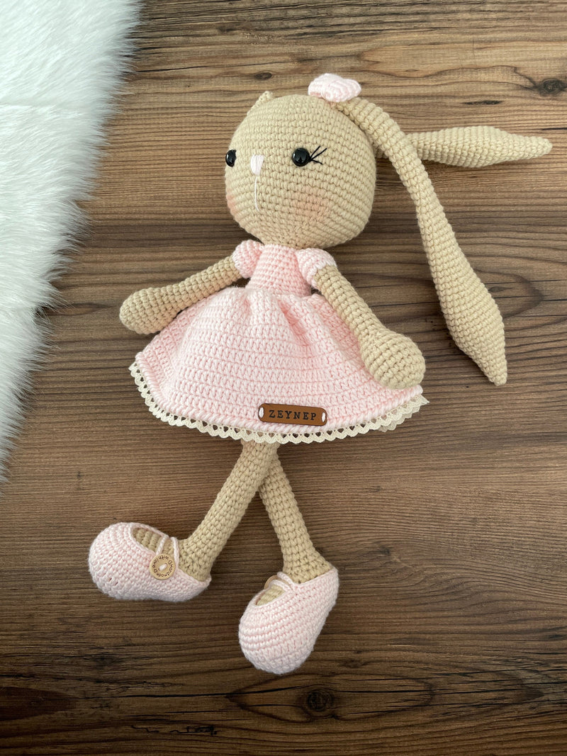 Handmade brown skin bunny rabbit toy crocheted using the amigurumi technique. Features a soft pink dress and a customizable name on a leather label. Perfect for baby gifts and nursery decor.