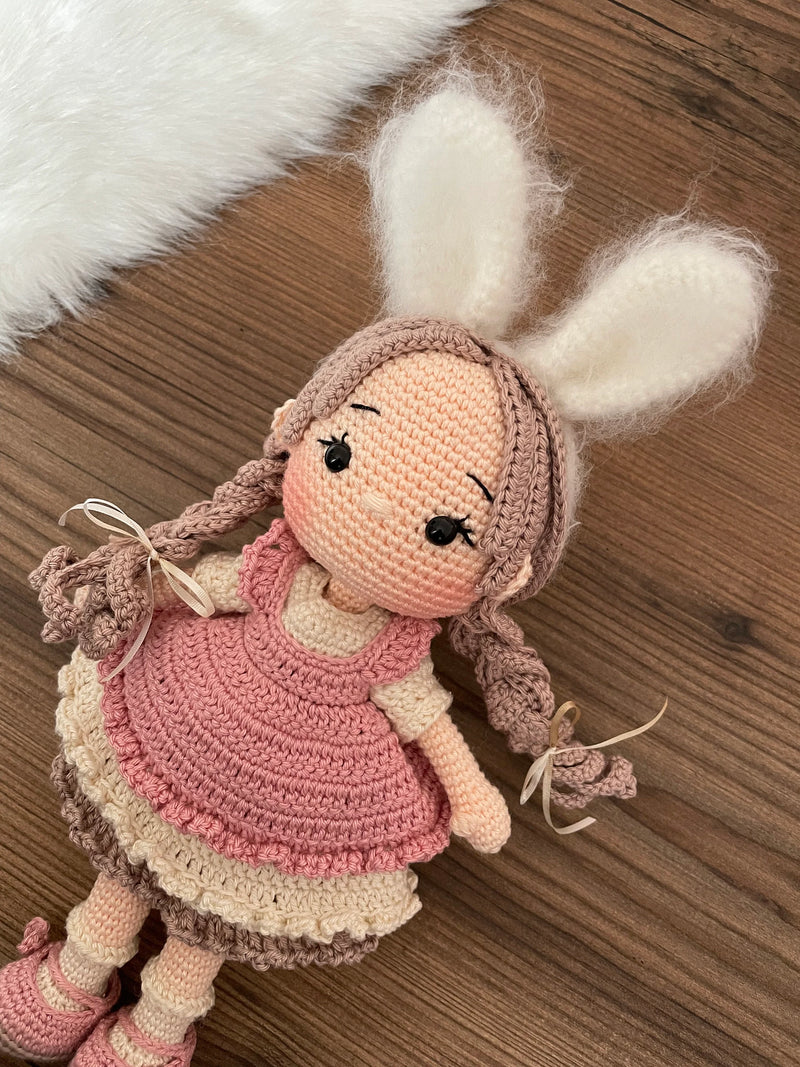 A handmade crocheted Rabbit Girl Doll with a pink dress, braided hair, and soft bunny ears. This amigurumi toy is customizable with a name on a leather label, making it a perfect choice for nursery decor and personalized gifts.