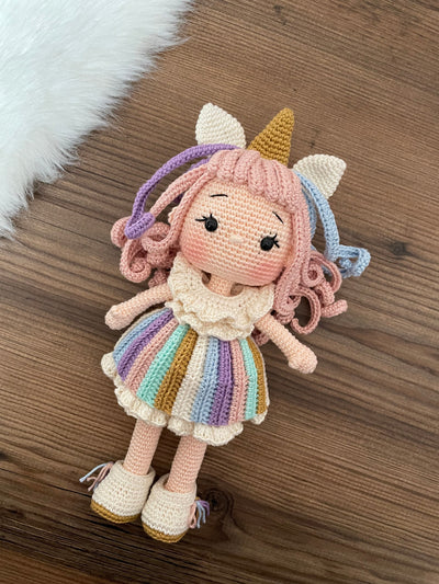 A handmade crocheted Unicorn Girl Doll with pastel rainbow dress, pink curly hair, and a golden horn. This amigurumi toy is customizable with a name on a leather label, perfect for nursery decor and personalized gifts.