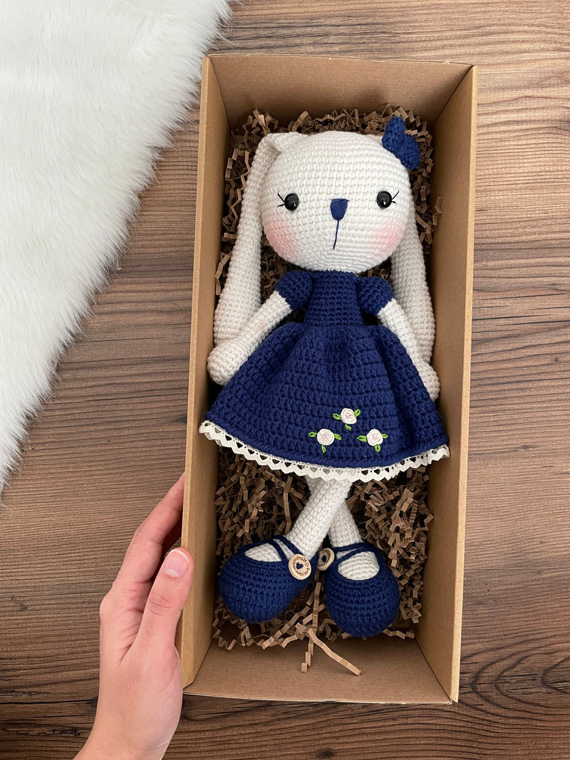 Handmade Navy Blue Dressed Bunny Rabbit Toy crocheted with amigurumi technique, featuring a cute navy blue dress with embroidered flower details and customizable name on a leather label. Lying inside a gift box. Perfect for kids and baby gifts.