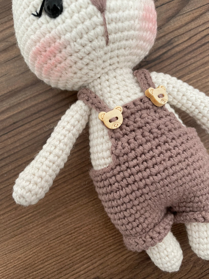 A mink color handmade crocheted Overall Girl Bunny Rabbit Toy with a cute bow and overalls. This amigurumi bunny doll is customizable with a name on a leather label, making it an ideal choice for nursery decor and personalized gifts.